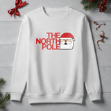 White sweatshirt with Santa Claus graphic and 'The North Pole' text on a wooden surface with Christmas decorations.