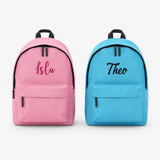 Two personalized backpacks shown side by side on a light gray background. The left backpack is pink with the name 'Isla' in dark pink script, and the right backpack is blue with the name 'Theo' in black script.