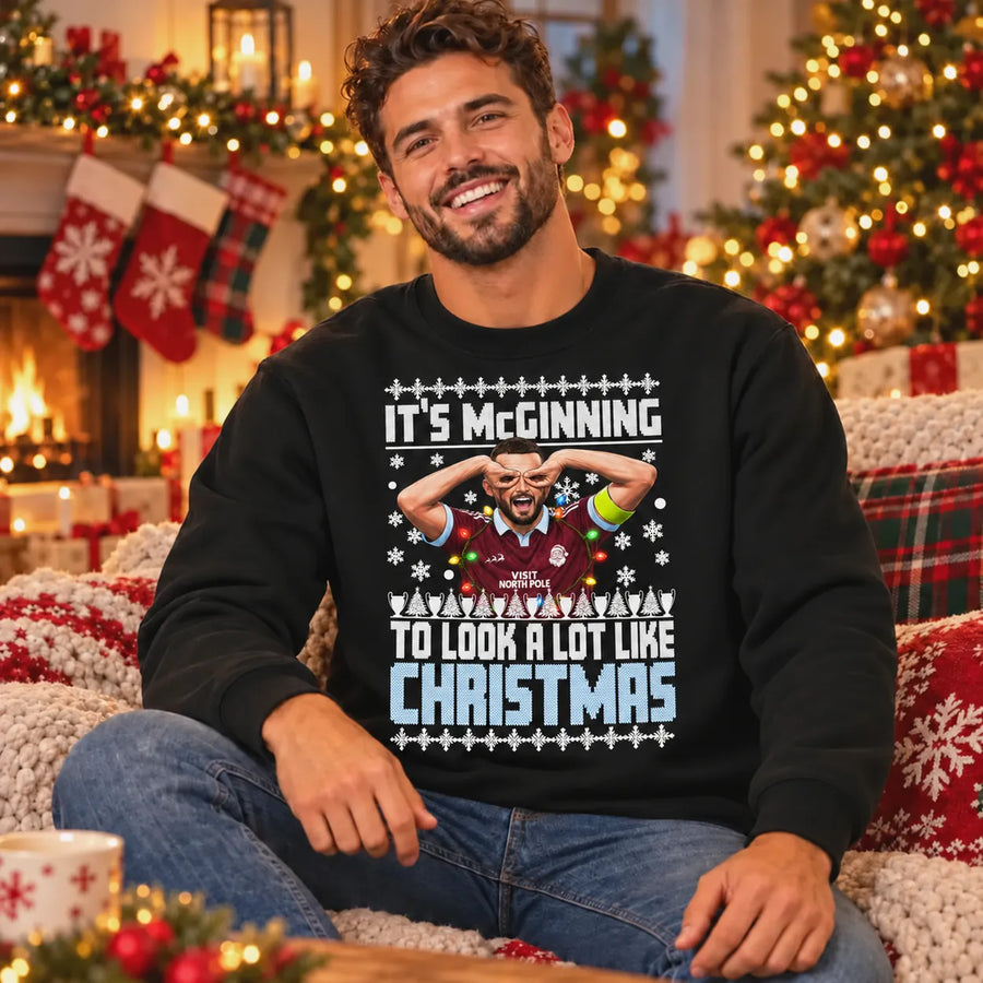 Male model wearing black John McGinn Christmas jumper with festive It's McGinning to Look a Lot Like Christmas print beside a decorated fireplace.